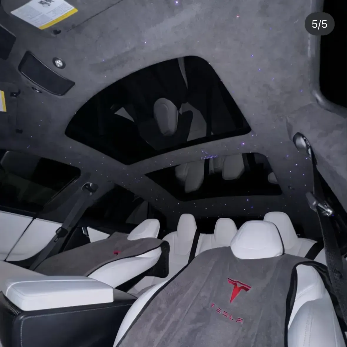 Interior of a Tesla with white seats, a panoramic roof, and starry ceiling lights. Gray seat covers with the Tesla logo are visible.