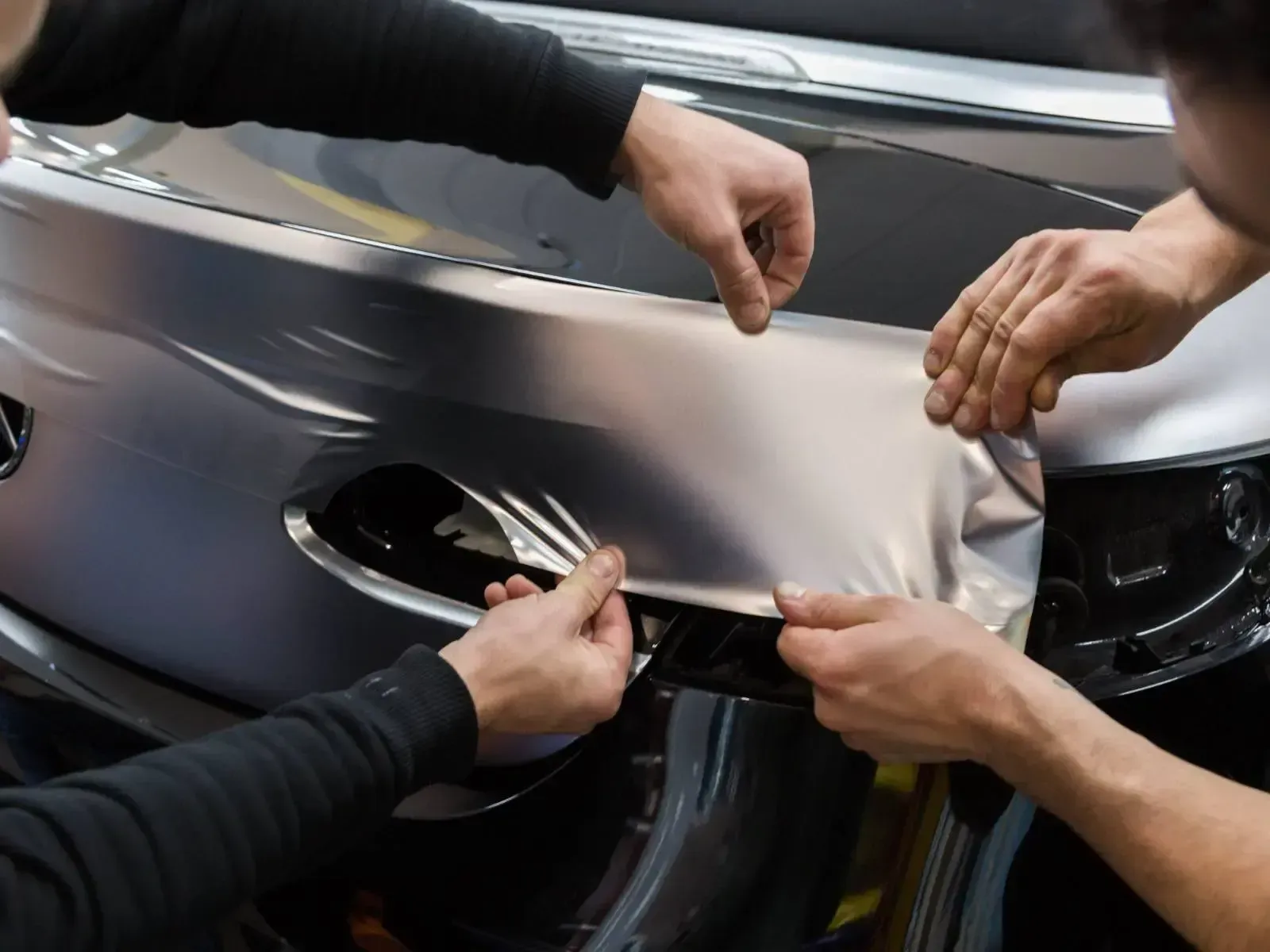 Two people applying matte grey vinyl wrap to a car bumper.