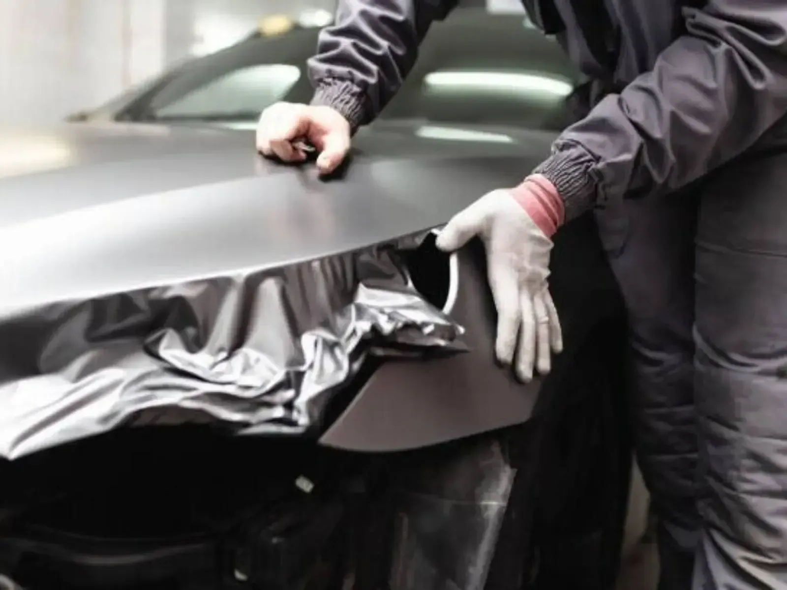 Person applying gray vinyl wrap to a car's front fender in a garage.