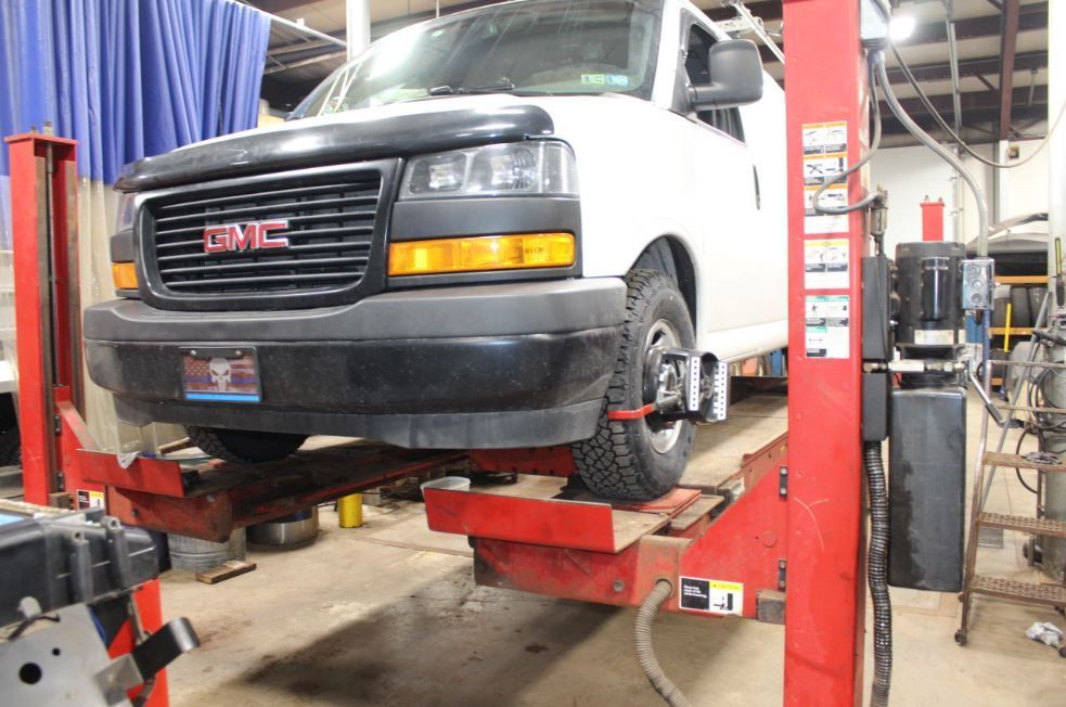 White GMC van on a red lift in a garage, undergoing a wheel alignment check.