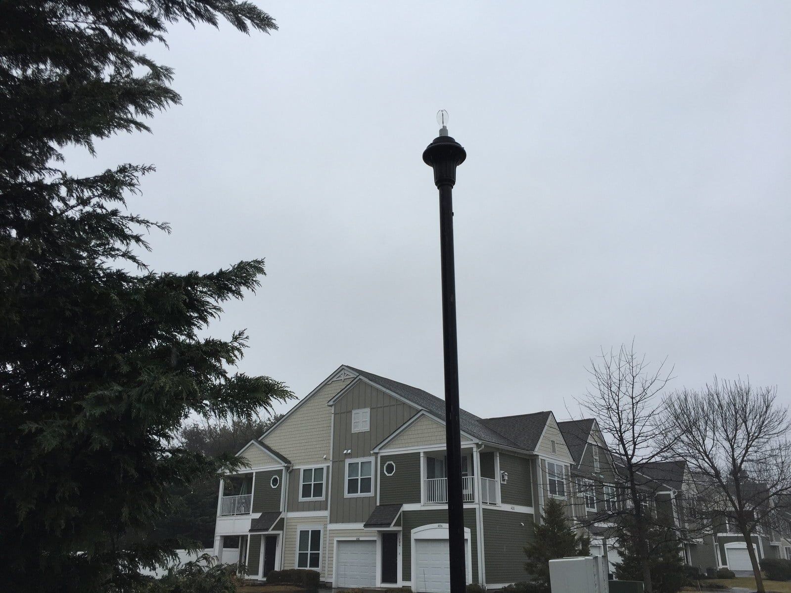Snowy residential street with a two-story house and a tall streetlamp under an overcast sky