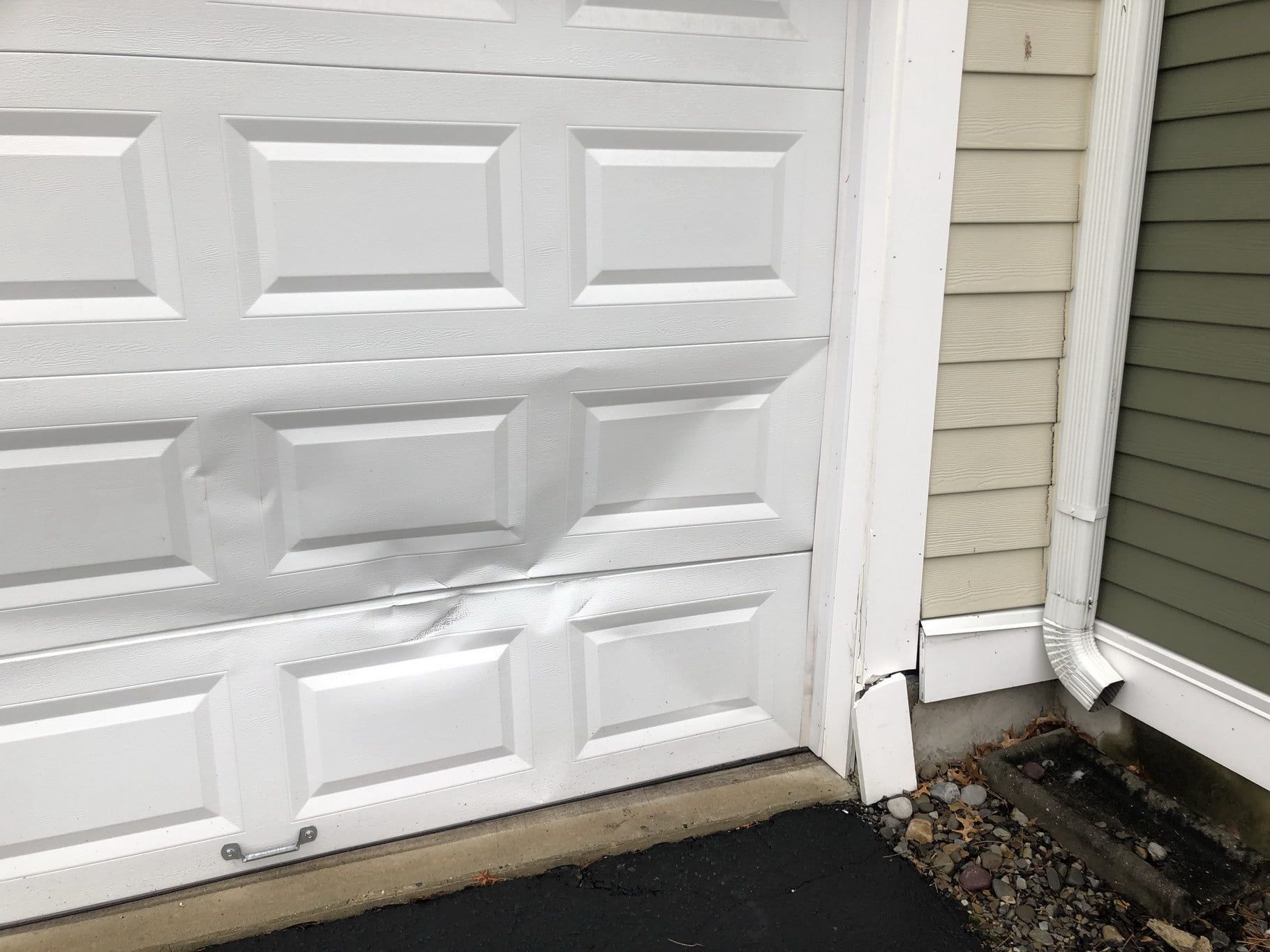 White garage door with a cracked lower right corner beside gray house siding and a paved driveway