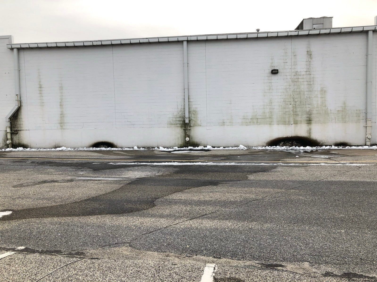 White industrial building wall with grime stains above an empty asphalt lot.
