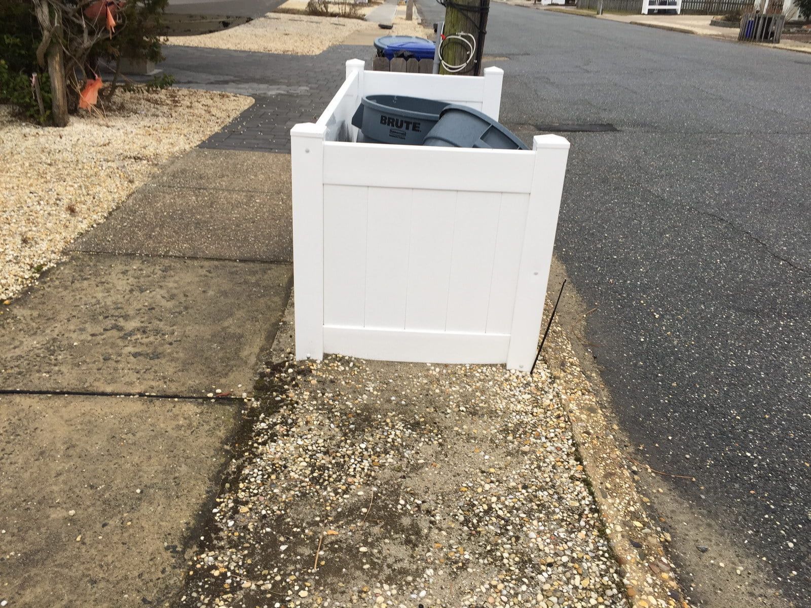 White trash bin on a sidewalk beside a residential street, filled with items and boxes.