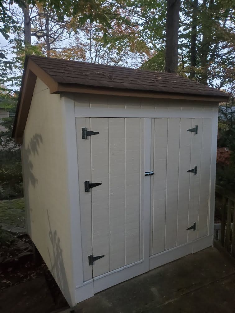 Small beige storage shed with white double doors and brown shingled roof outdoors among trees