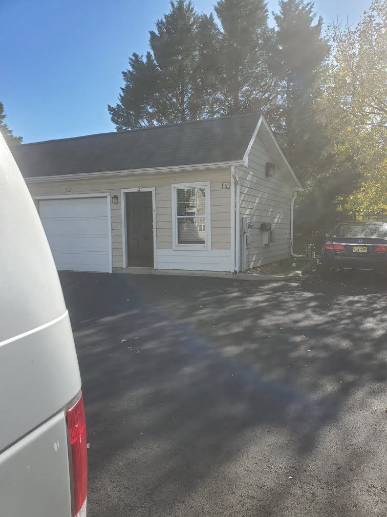 Small white house with a garage, driveway, and a parked vehicle in bright sunlight.