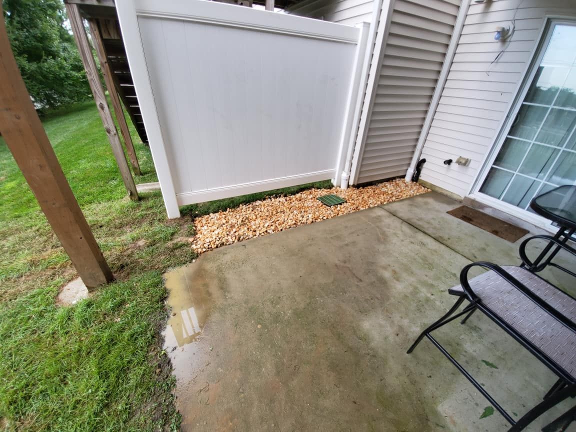 Back patio with wet concrete, white fence, mulch strip, and a chair beside the house.