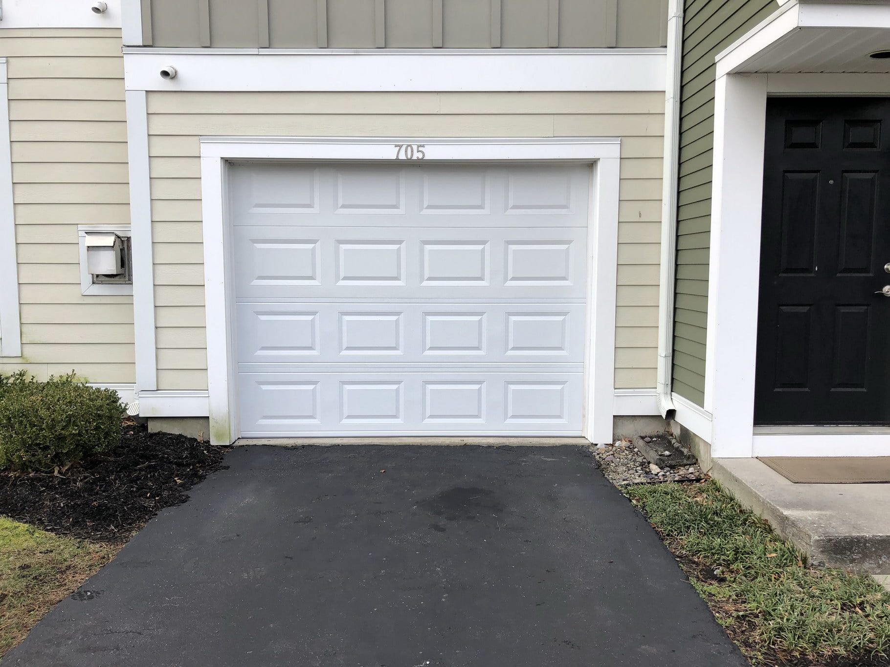 White garage door with a driveway at a townhouse entrance, beside beige siding and a black front door.