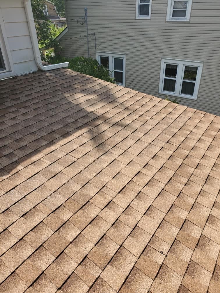Brown shingle roof viewed from above beside a beige house with windows and a small tree shadow