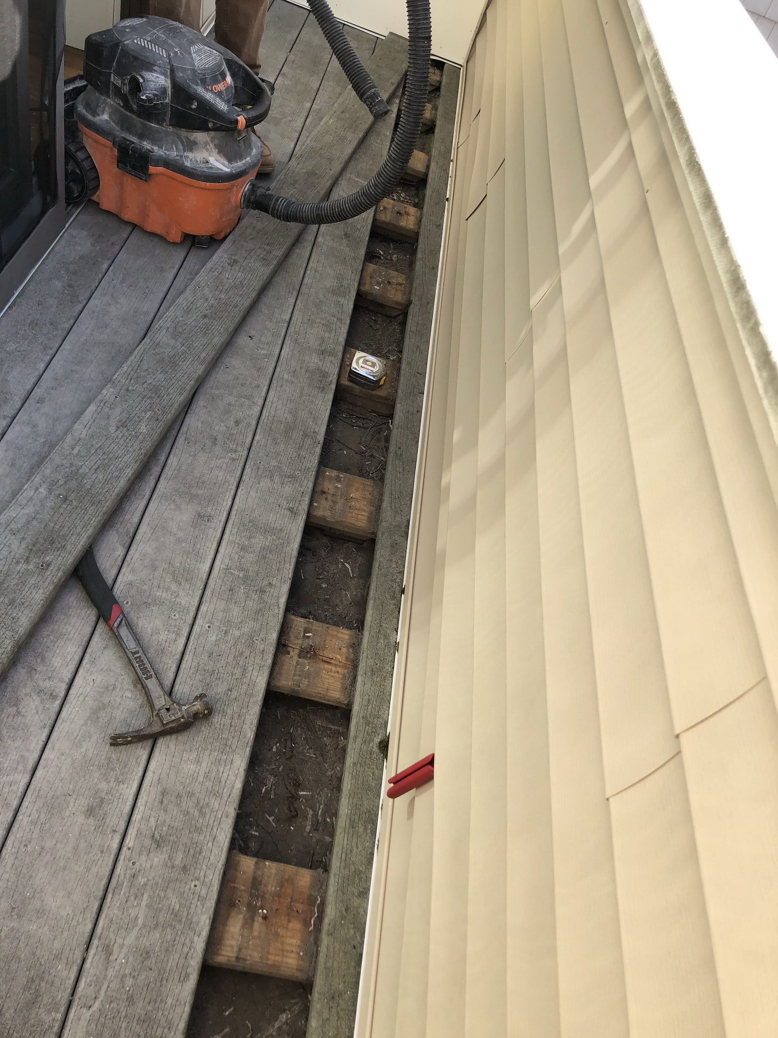 Rooftop gutter gap beside beige siding, with exposed wooden supports and tools on the roof.