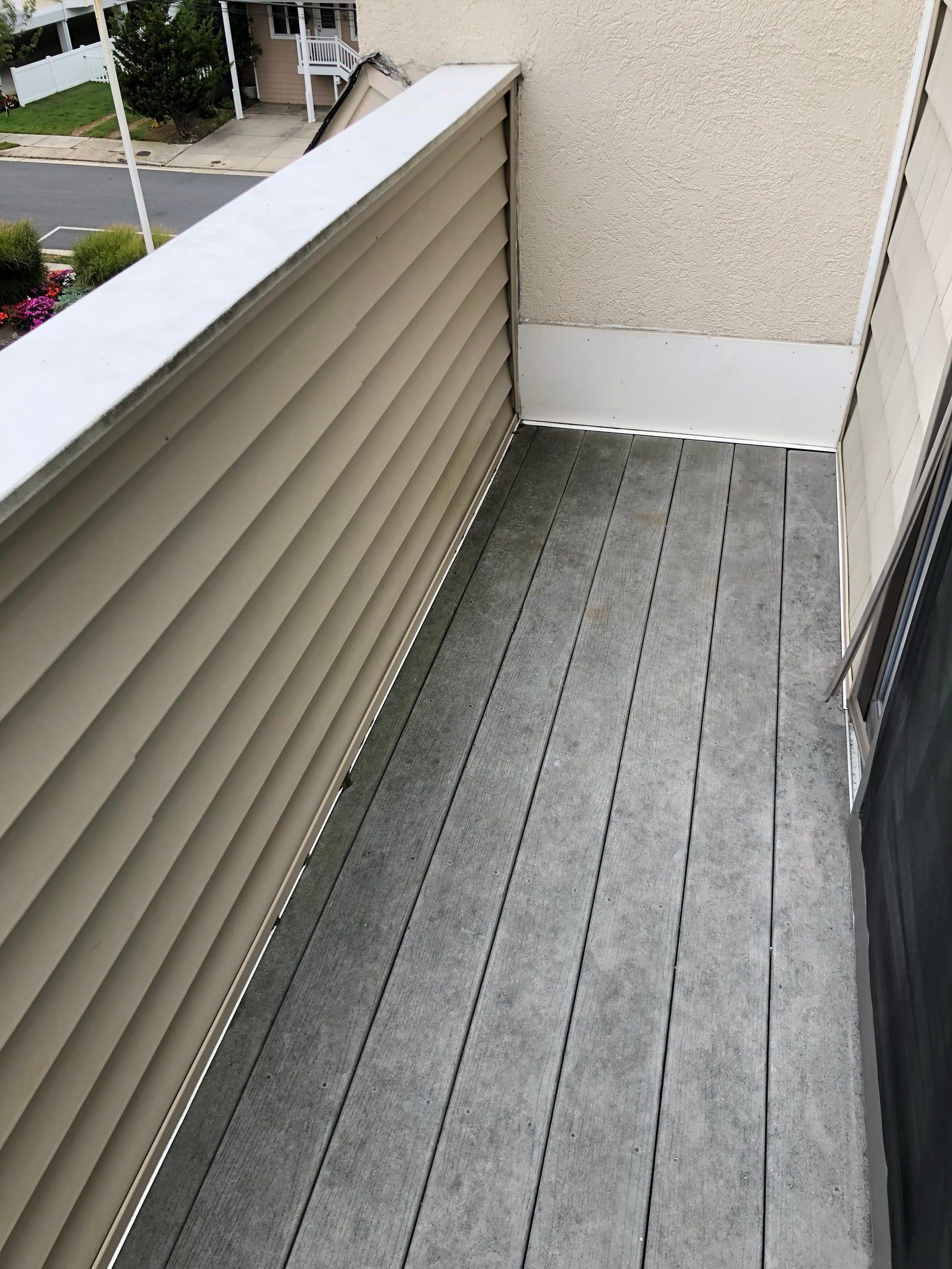 Narrow apartment balcony with gray flooring and beige railing, overlooking an outdoor street.