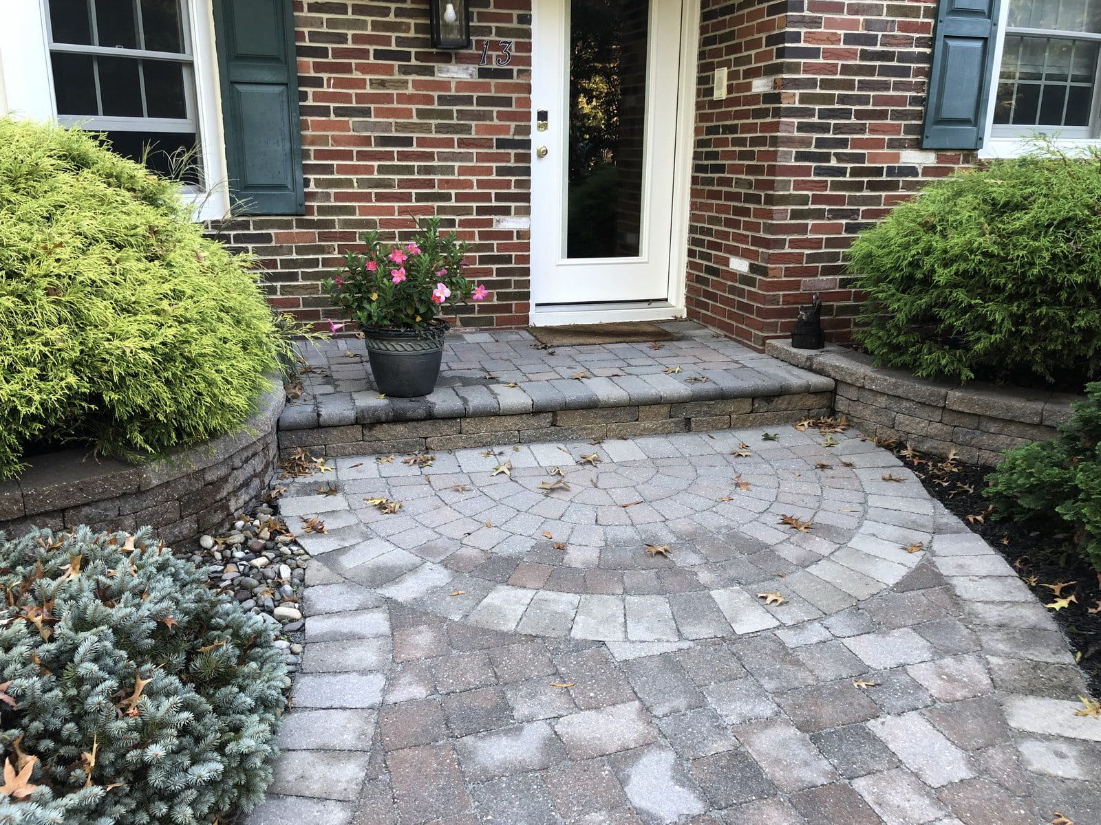 Front walkway with curved stone pavers leading to a brick house entrance, bordered by shrubs and flowers
