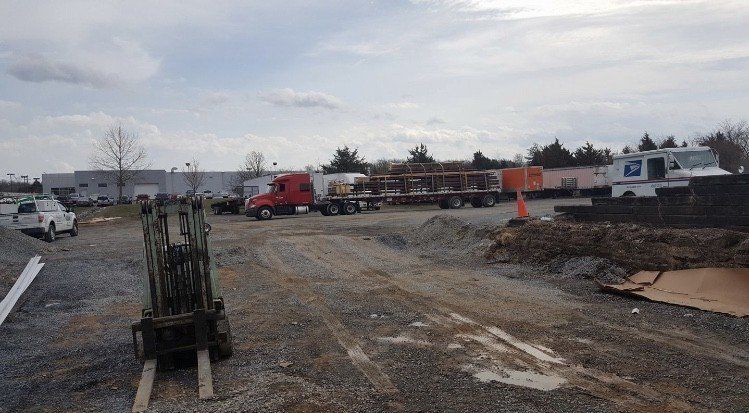 Construction site: Red truck, flatbed with wood, backhoe in foreground, cloudy sky.