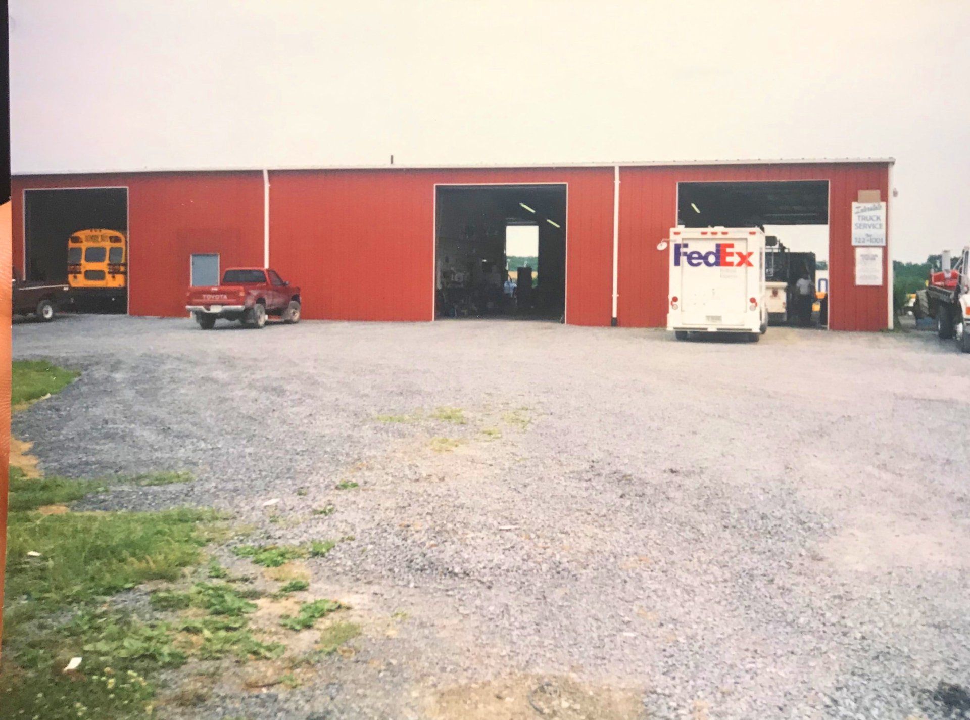 Red building with open garage doors; vehicles, including a school bus and FedEx truck, are visible. Gravel lot.