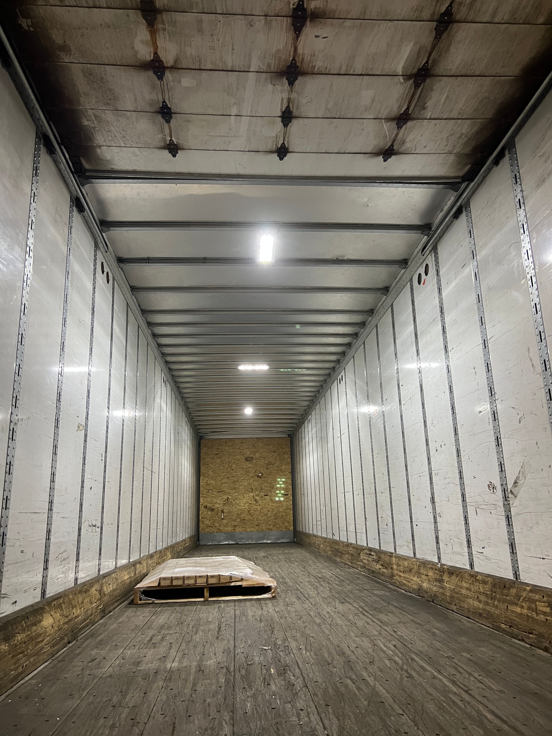 Inside a dirty cargo trailer, a pallet sits near the back. Walls and ceiling are light, with metal reinforcement.