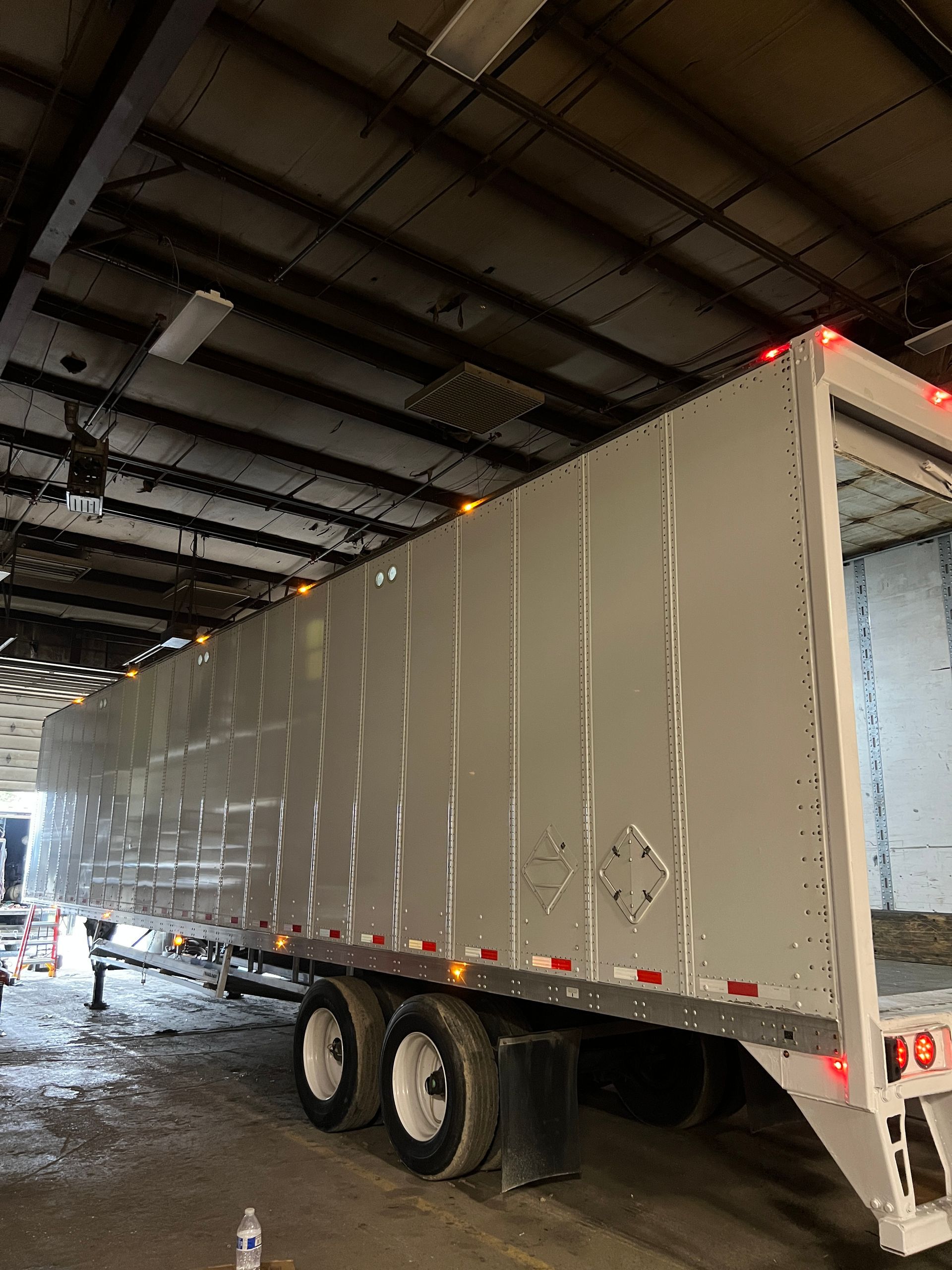 Silver semi-trailer in a dim warehouse, illuminated by overhead lights. Open door reveals cargo space.