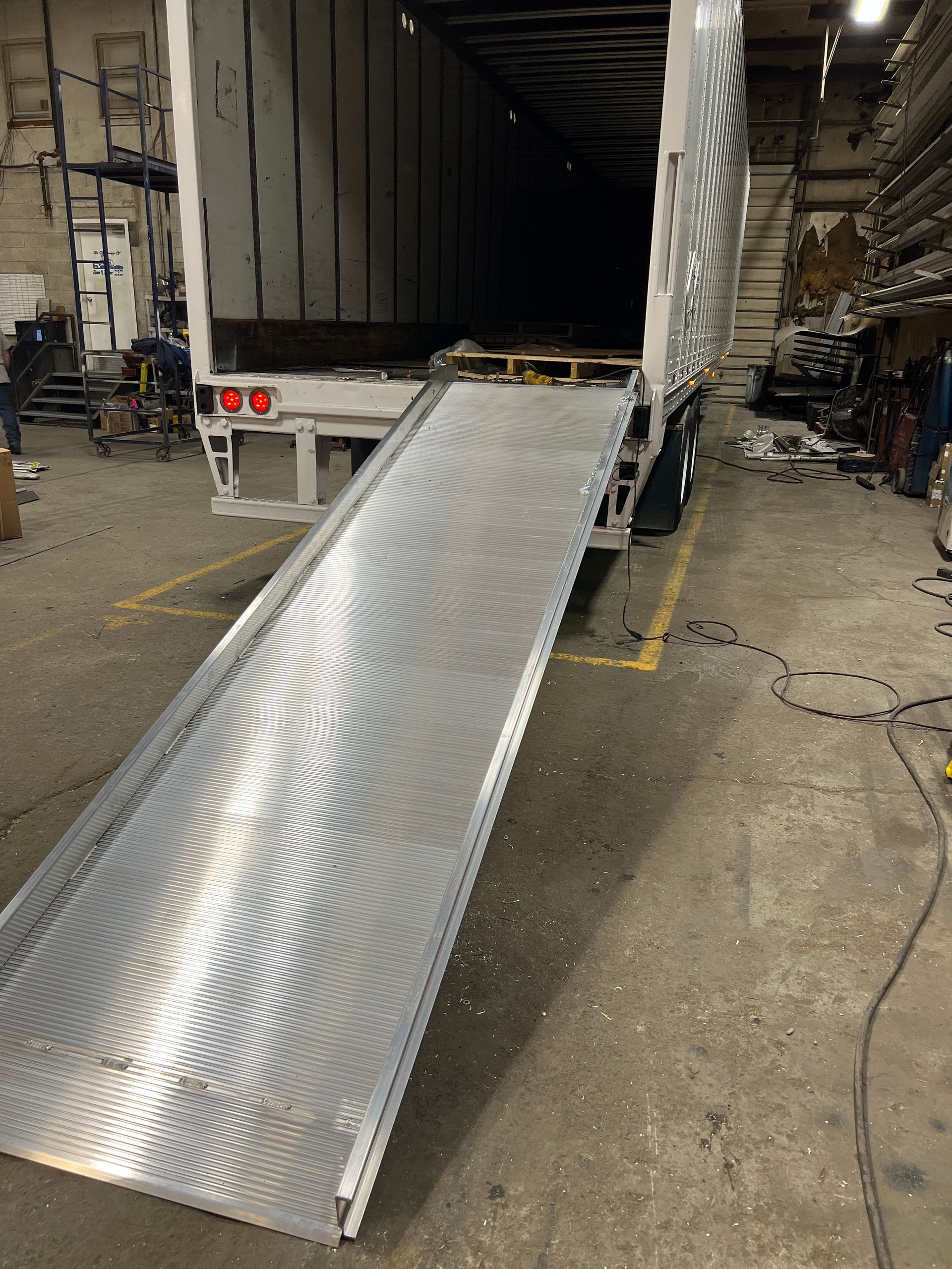 A metal loading ramp extends from an open truck trailer, on a warehouse floor.