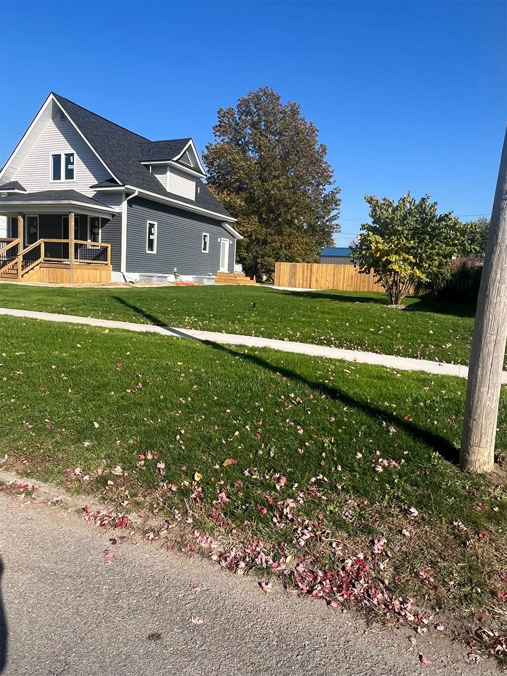 Gray house with porch and black roof on a green lawn, sidewalk and fence in the background, under blue sky.