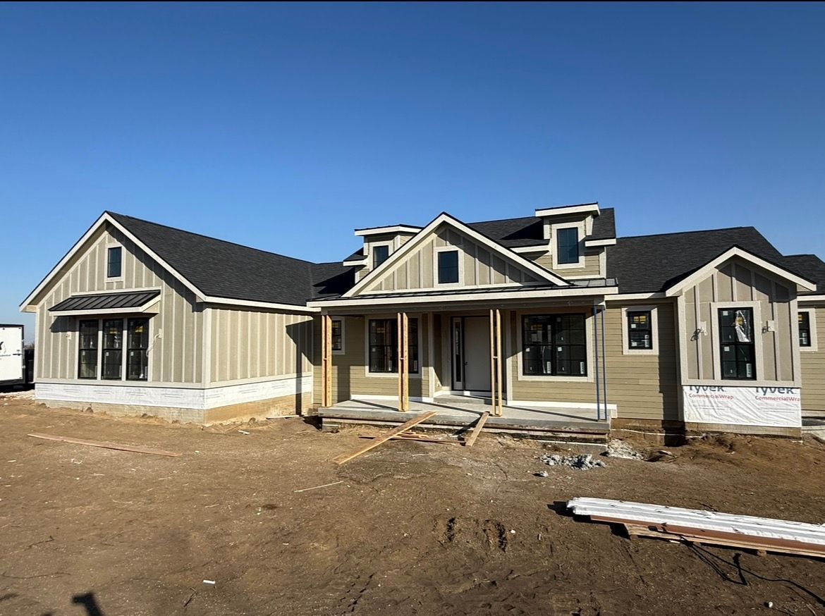 A single-story, neutral-toned house under construction with board-and-batten siding and a dark roof under a clear blue sky.