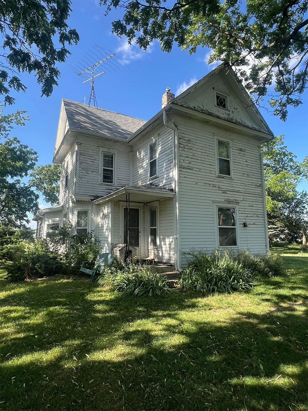White two-story house with gabled roof, overgrown foliage, and a blue sky in the background.