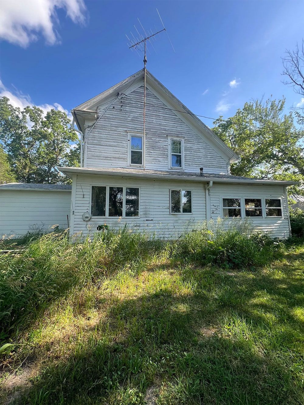 Dilapidated white two-story house with overgrown weeds, and an old antenna against a blue sky.