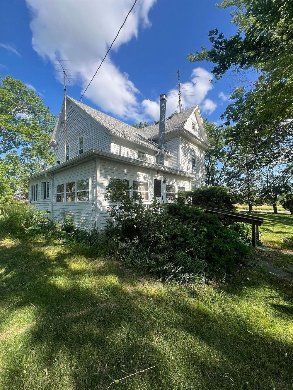White, two-story house with gabled roof, surrounded by trees and overgrown bushes on a sunny day.