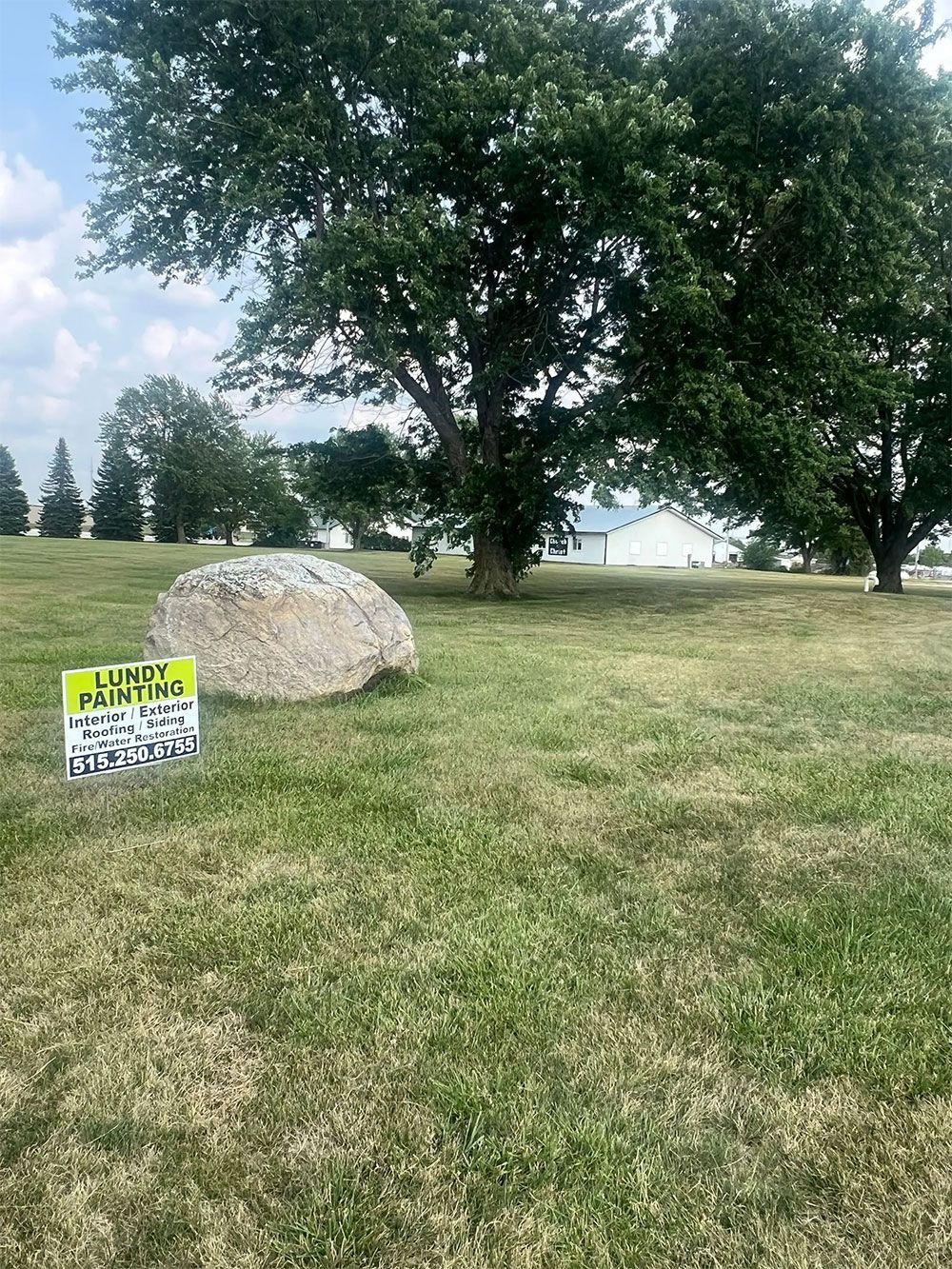 Large rock in a grassy field with trees. Sign posted in the grass.