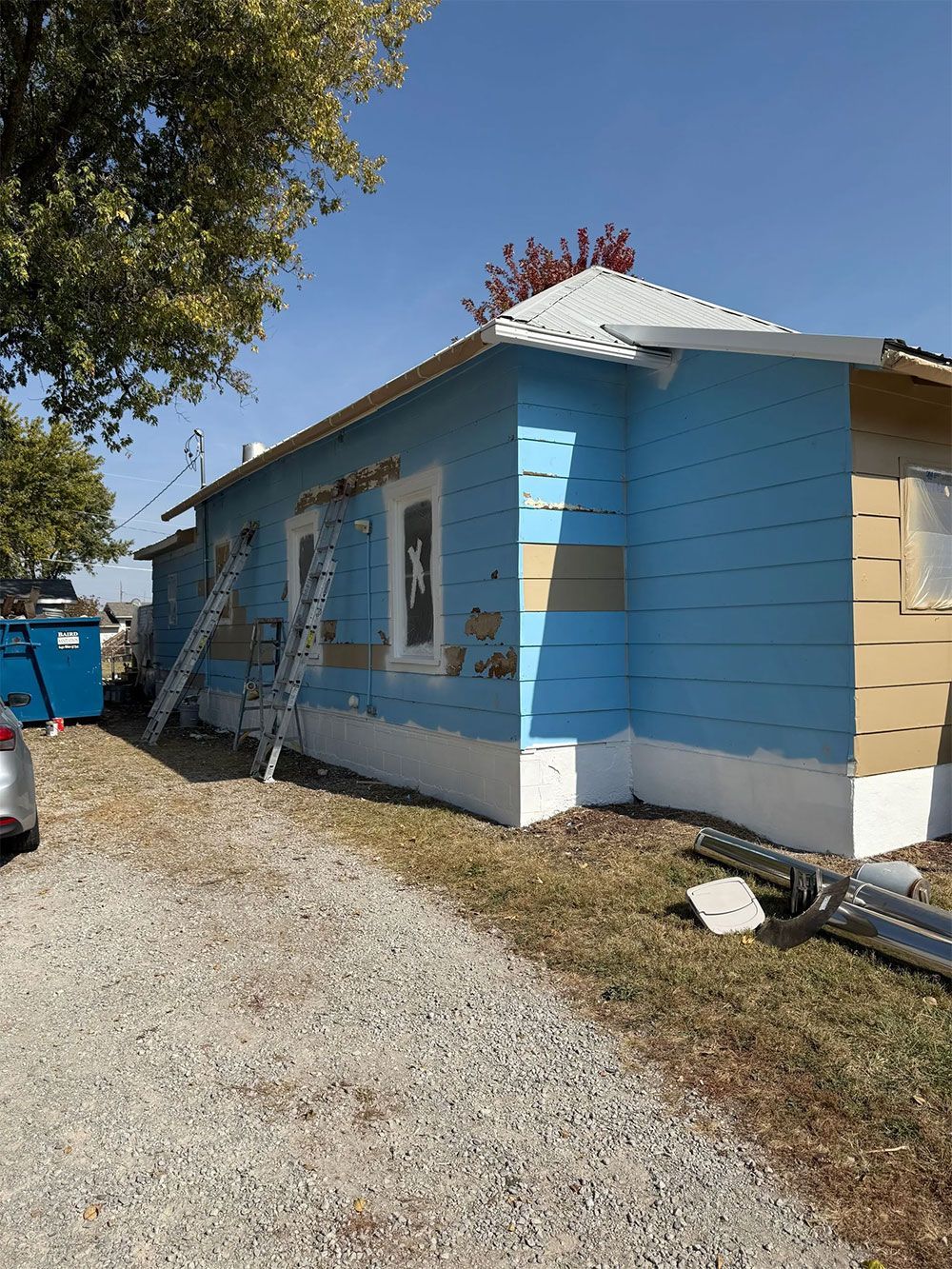 House exterior being painted, blue siding, ladders, partially completed, blue dumpster, clear sky.