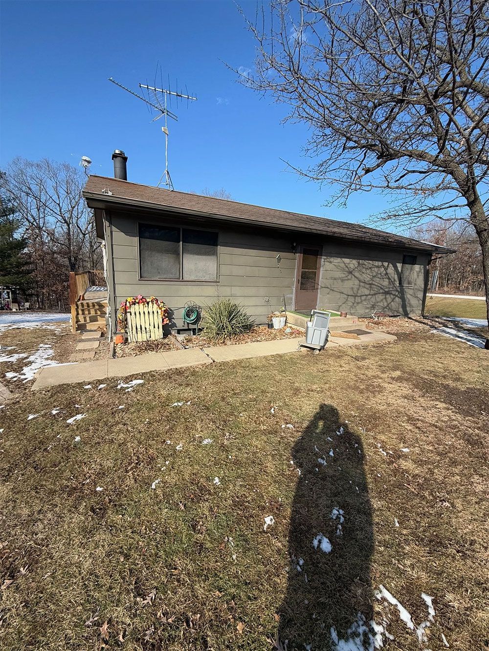 Small, weathered, one-story building with slanted roof, antenna, and chimney. Bare trees and patches of snow on the ground.