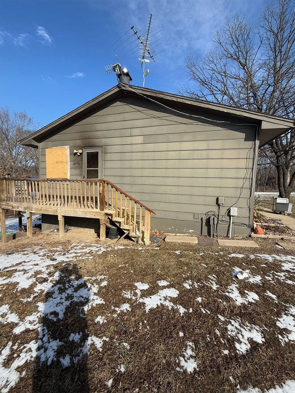 Exterior of a house with damaged siding, boarded window, deck, and snow on the ground.