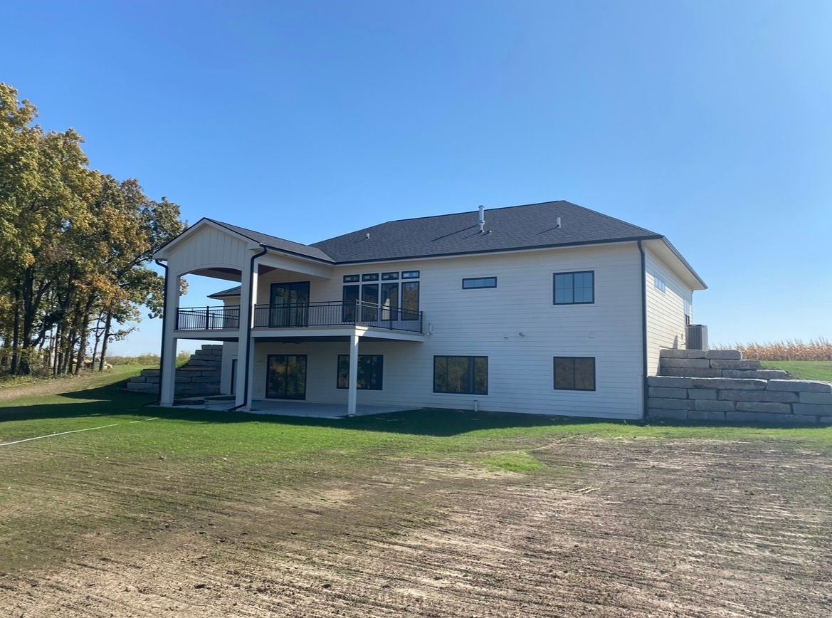 A two-story light-colored house with a dark roof and a rear balcony, situated on a grassy lot near a field and trees.