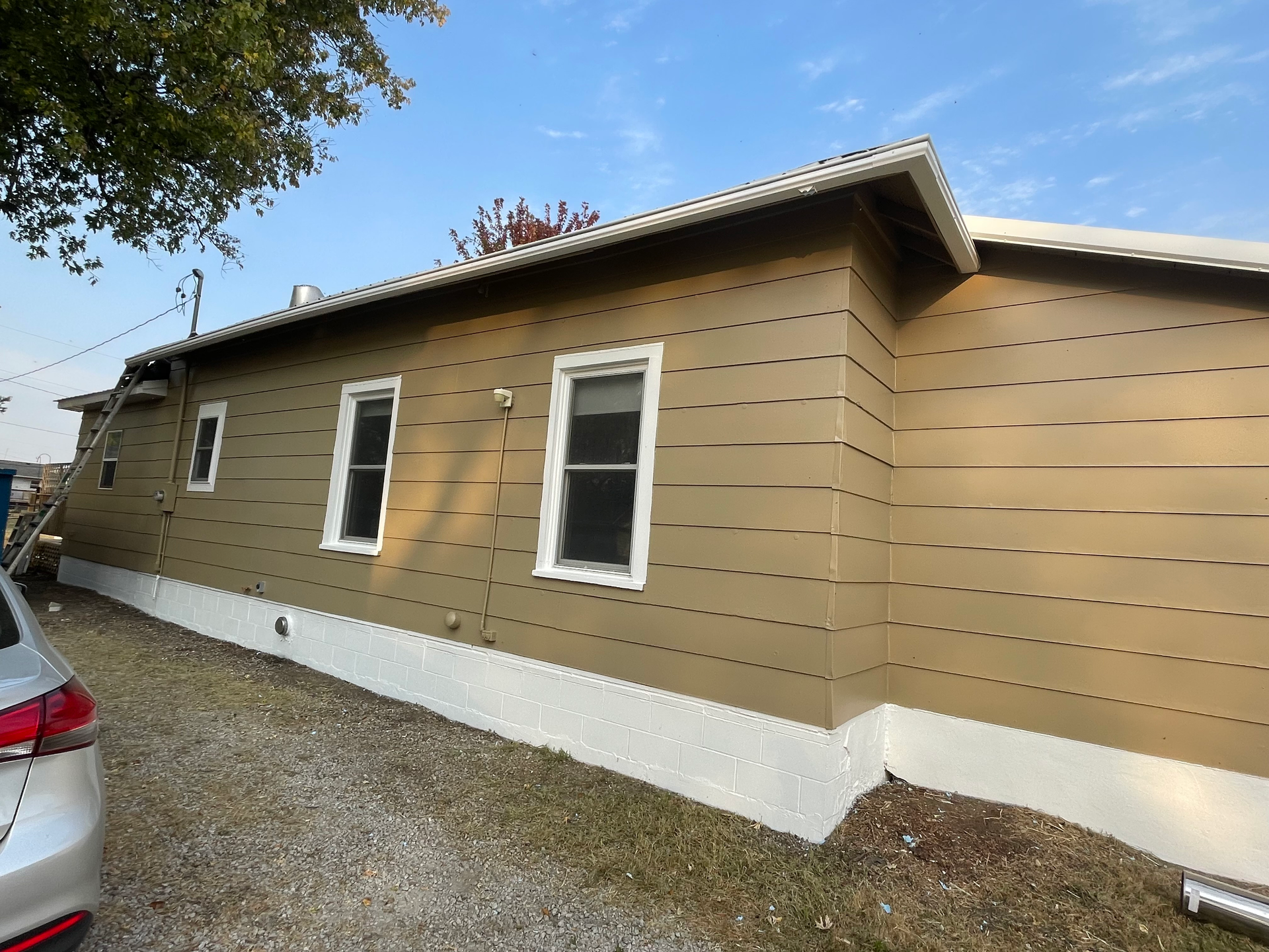 Tan house with white trim, windows, and foundation against a blue sky; car parked to the left.