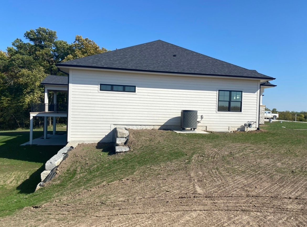 A side view of a white, modern house with a dark gray hip roof, set on a grassy hill with a small stone retaining wall.