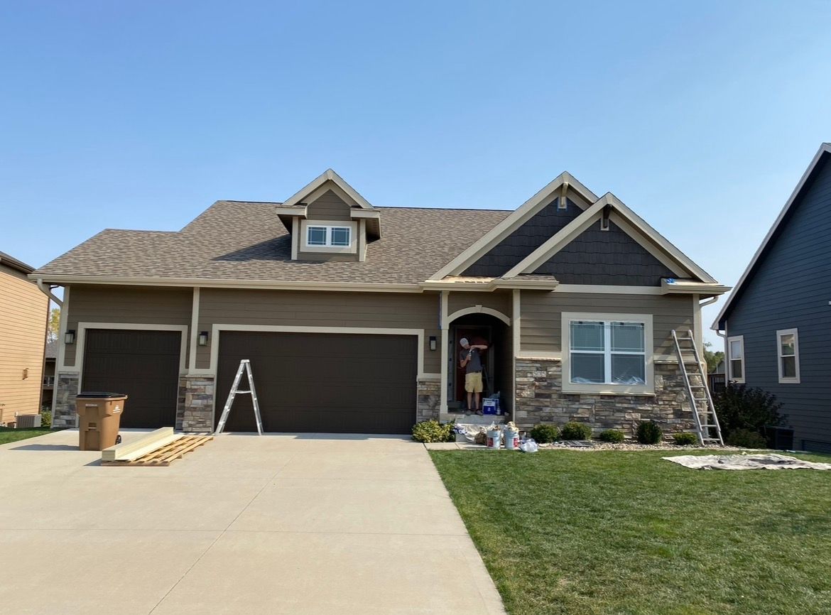 A one-story tan and brown suburban house with a multi-car garage, stone exterior accents, and ladders on the driveway.