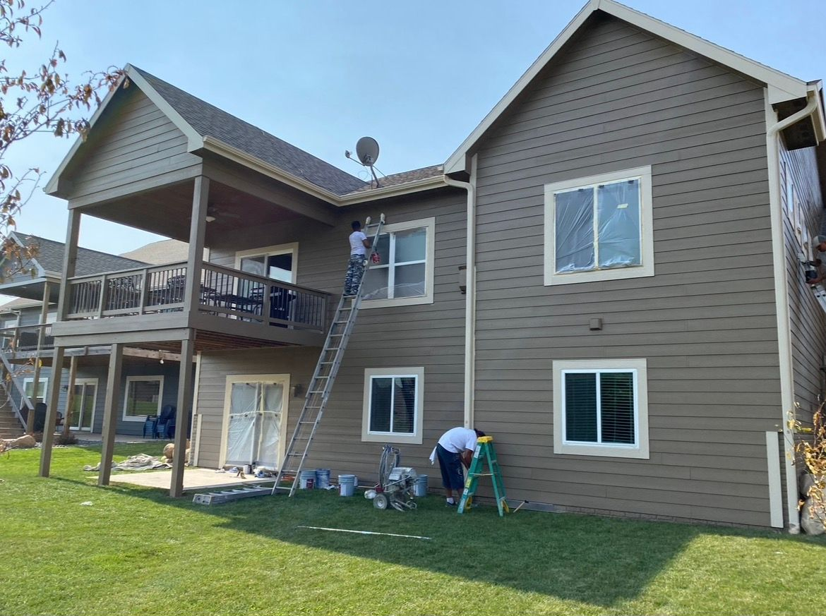 Two workers paint the exterior of a brown house; one uses a tall ladder, while the other uses a step ladder.