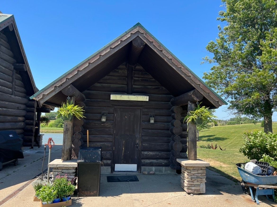 A rustic dark-wood cabin with a gabled roof, stone-based pillars, potted plants, and an entrance door under a sunny sky.