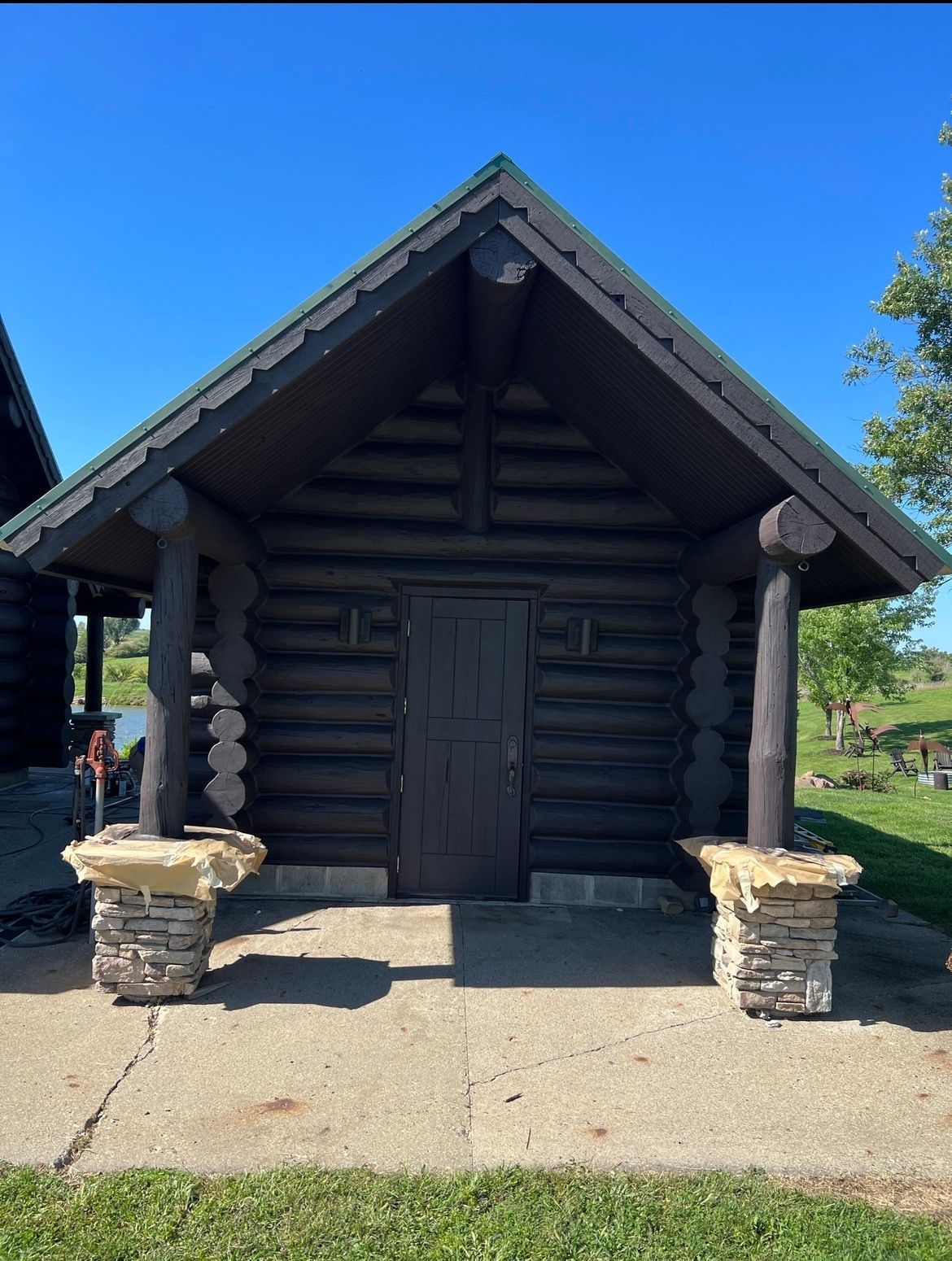 Dark brown log cabin with a peaked roof, supported by two stone-based pillars, standing on a concrete porch under blue sky.