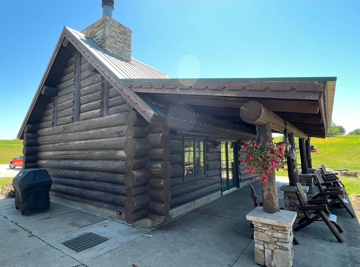 A rustic, dark-wood log cabin with a stone chimney, a covered porch with stone-based pillars, and flowers on a sunny day.