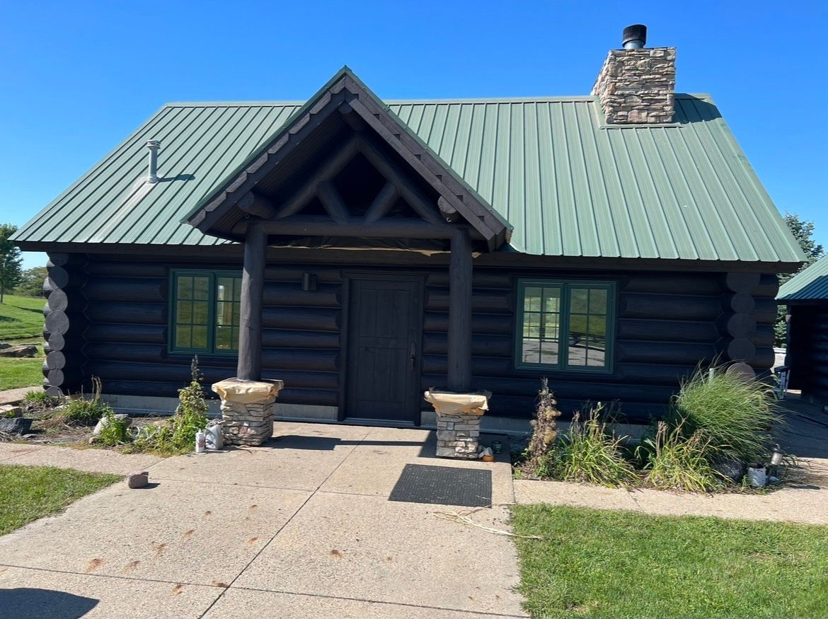 A dark wood log cabin with a green metal roof, a stone chimney, and a central porch on a clear, sunny day.