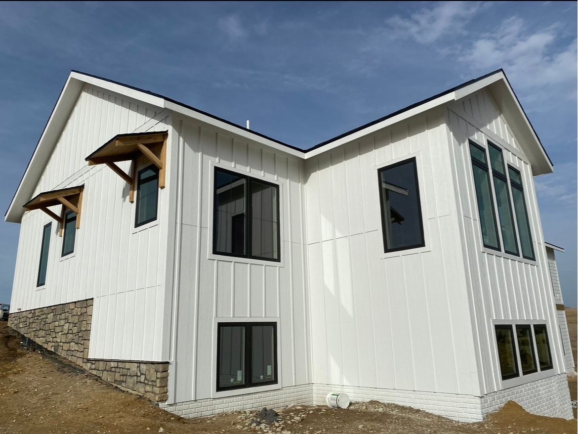 A white, modern farmhouse with board-and-batten siding, black window frames, and stone accents on a sloped dirt lot.