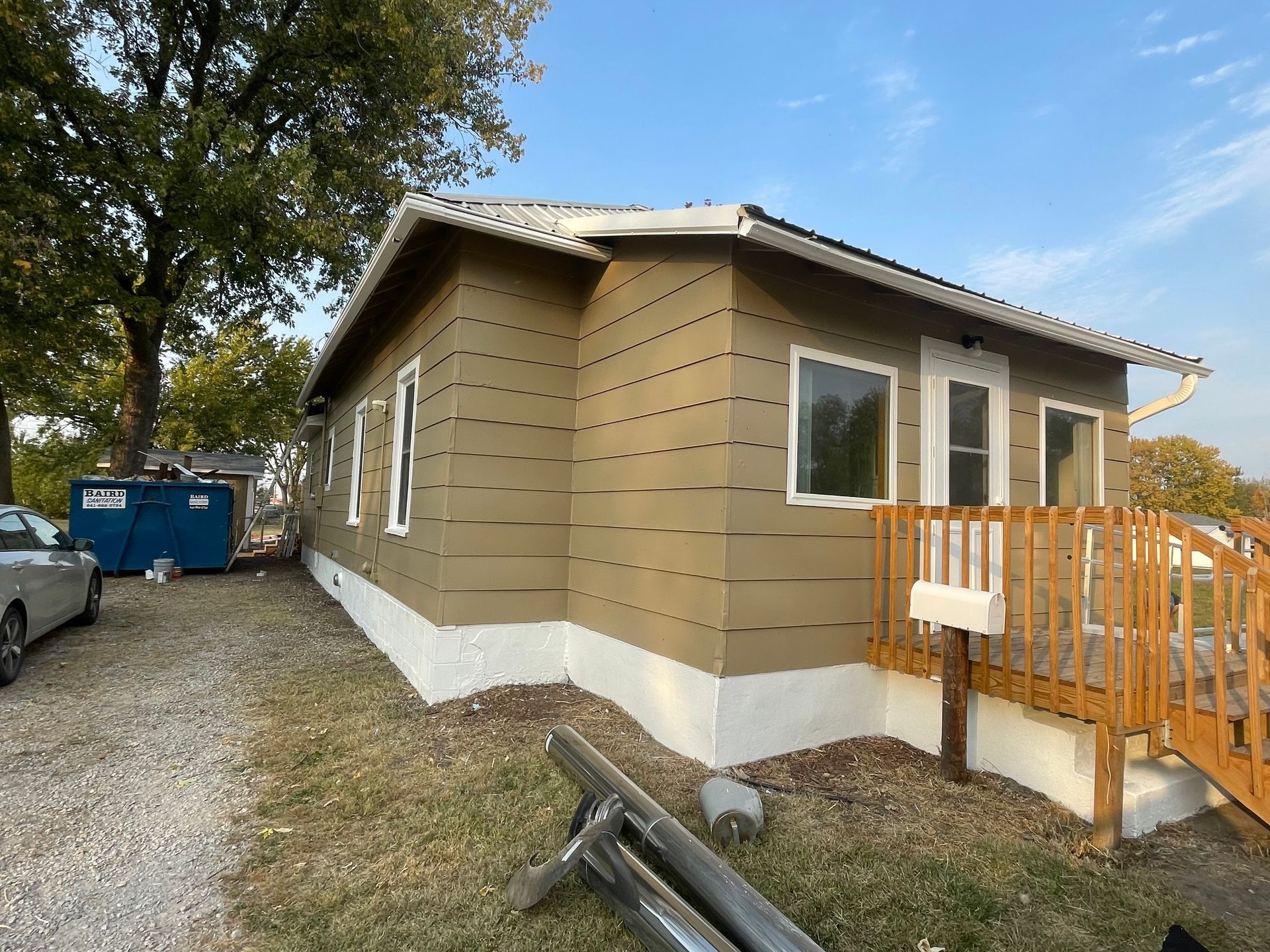 Tan house with white trim, wooden deck, blue dumpster, and gravel driveway.