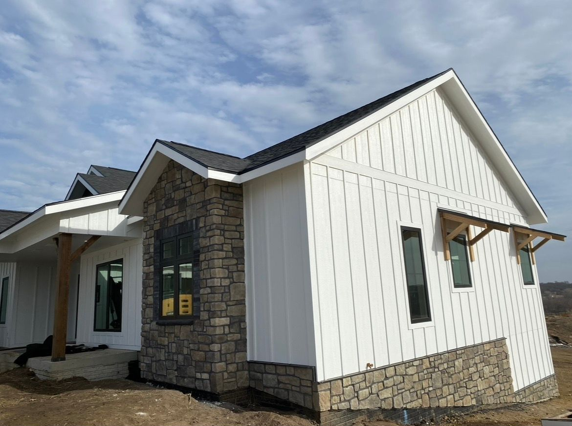 A new house with white board-and-batten siding, stone accents, and a dark roof under a cloudy blue sky.