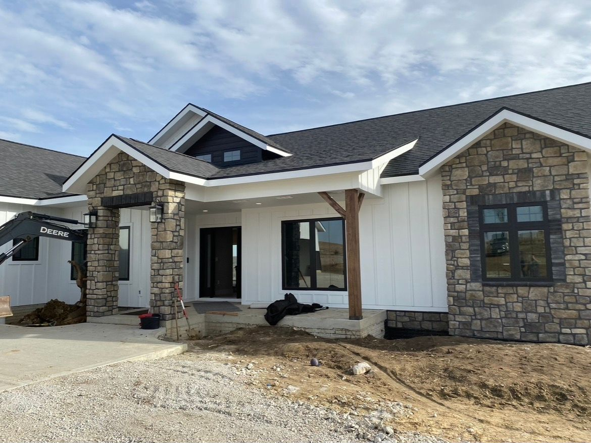A single-story house under construction featuring white board-and-batten siding, stone veneer accents, and a dark roof.