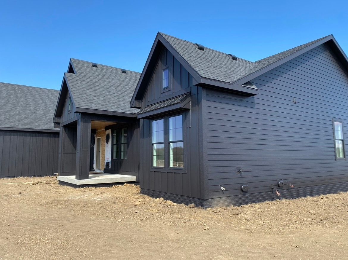 A new dark-painted, gabled residential home under construction against a clear blue sky, set on a gravel lot.