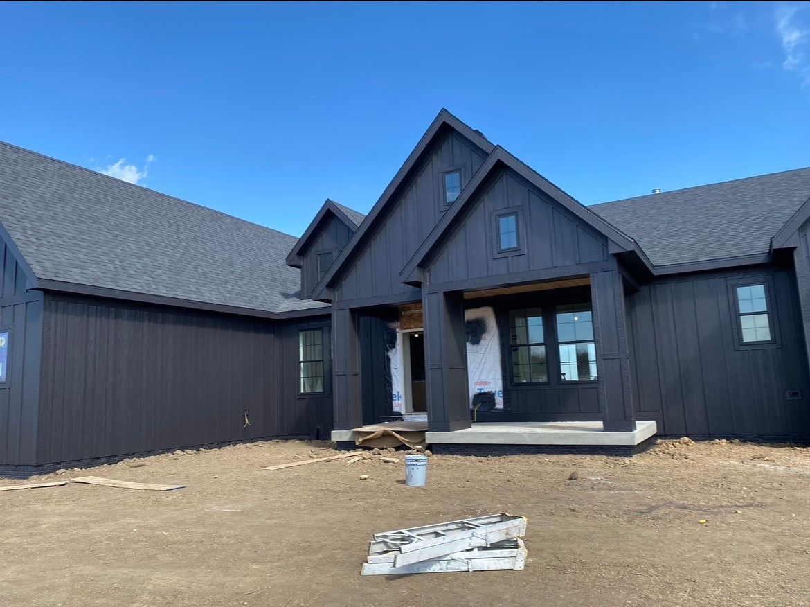 A modern, dark-sided farmhouse with a prominent front entryway, gray roof, and unfinished landscaping under a blue sky.