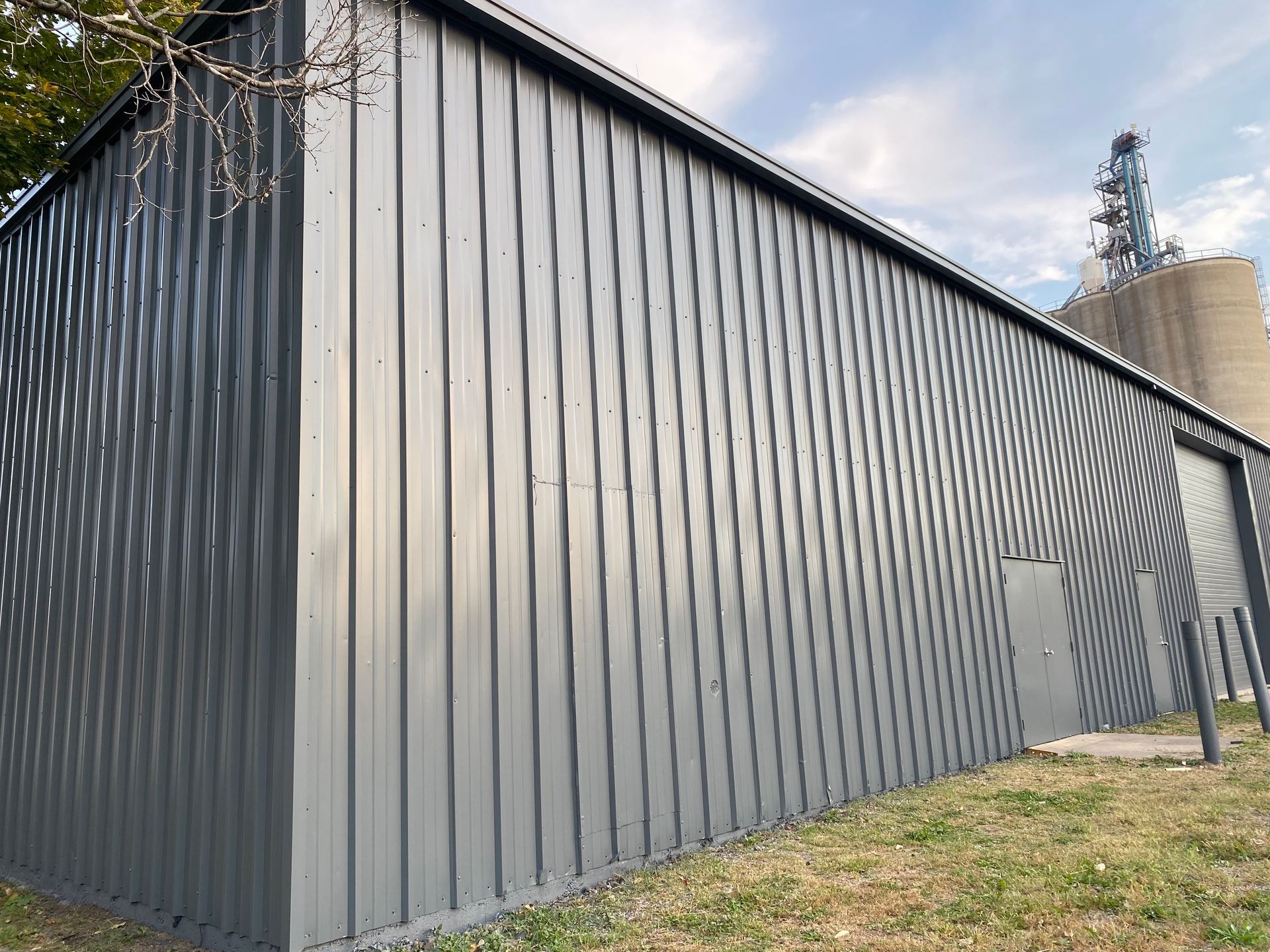 Gray metal building with vertical grooves, set on a grassy area, with silos in the background.