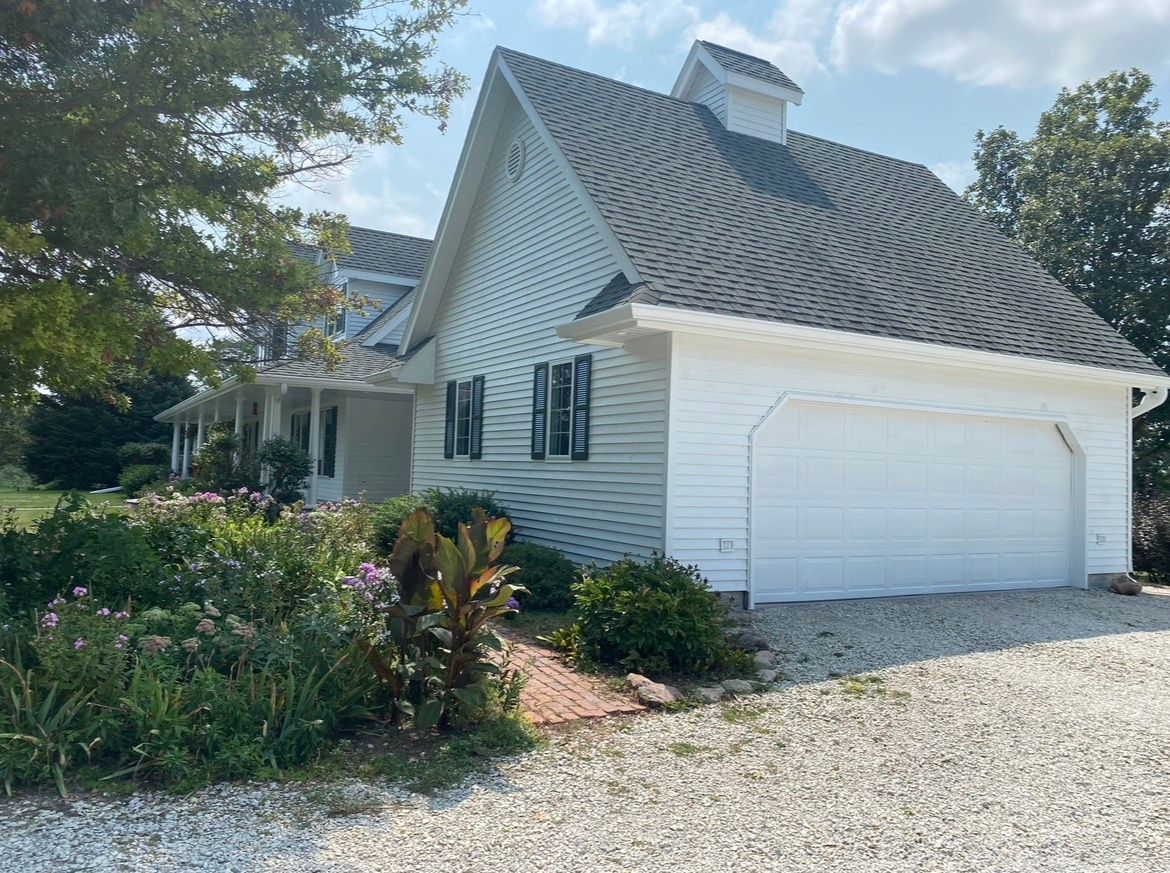 A white house with a garage and a cupola on its gabled roof, surrounded by trees and a flower bed on a gravel driveway.