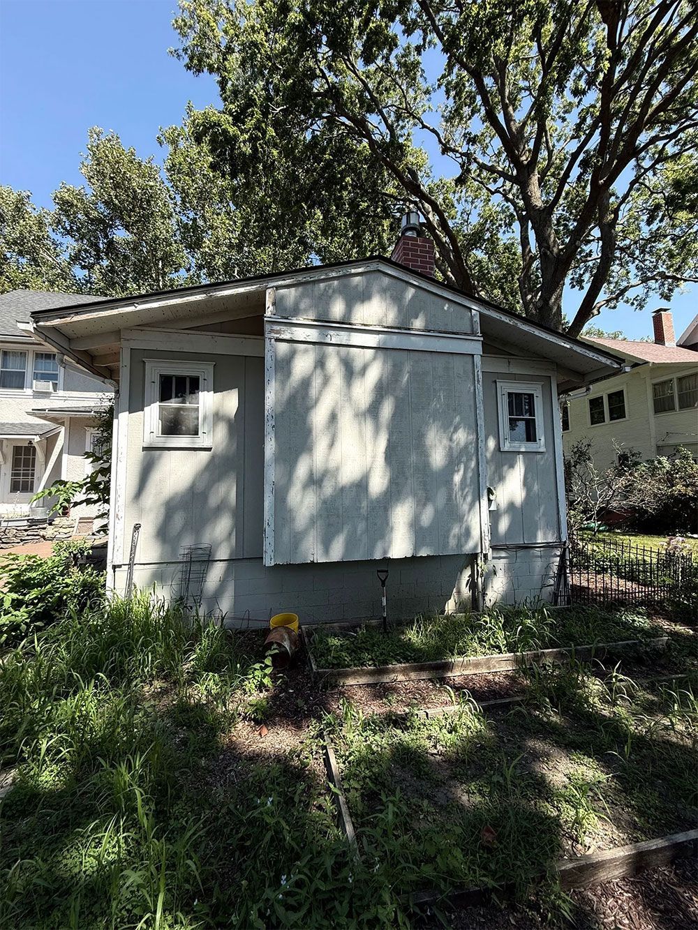 Small, weathered gray building with two windows. Overgrown weeds in foreground, large tree in background.