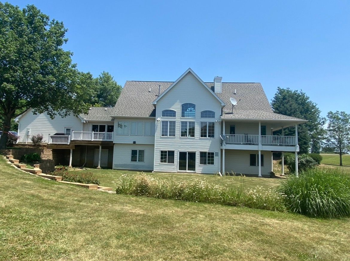 A large two-story white house with a wraparound porch and gray shingled roof, set in a sunny, grassy landscape.