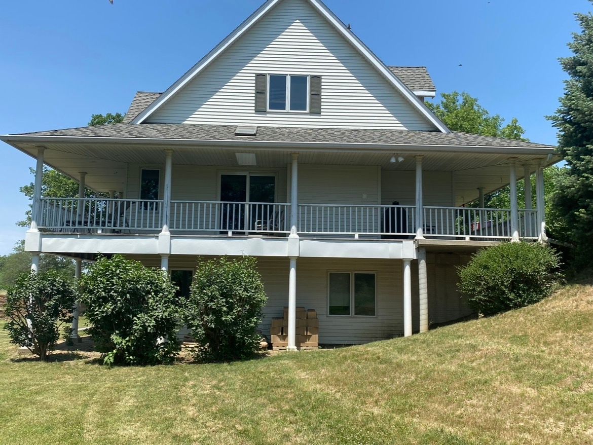 A two-story house with a white wrap-around deck, light-colored siding, and a gabled roof set on a grassy, sloped lot.